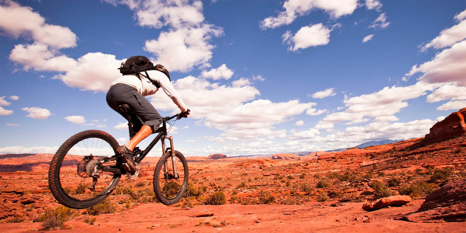Mountain biker in mid-air performing jump over red desert landscape with rocky terrain, sparse vegetation, and dramatic blue sky filled with white clouds overhead.