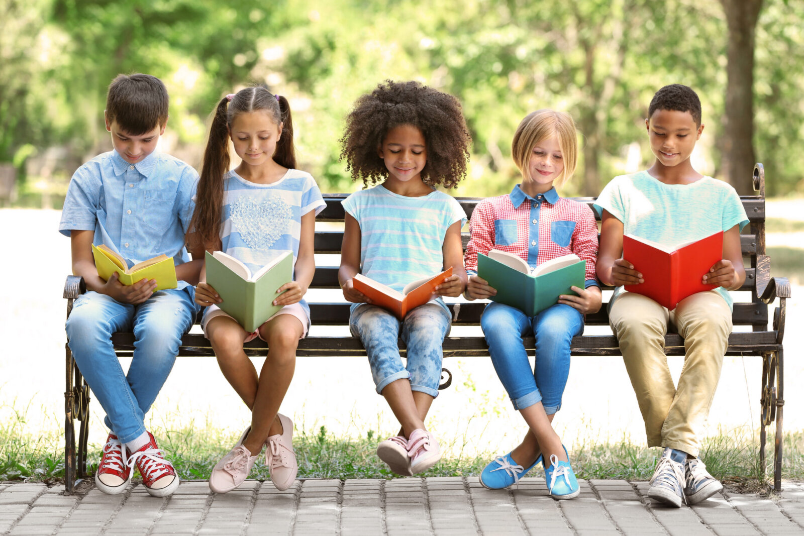 Five children of different ethnicities sit on a wooden park bench reading colorful books, surrounded by green trees and dappled sunlight on a peaceful day.