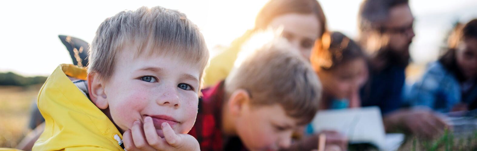 Young blonde child in bright yellow jacket smiling at camera while resting chin on hand, with family members gathered around table in soft golden sunset lighting.