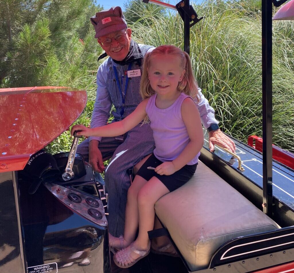 A smiling train conductor in striped overalls poses with a happy young girl driving a miniature train at Settlers Point RV Park in Utah, surrounded by lush greenery.