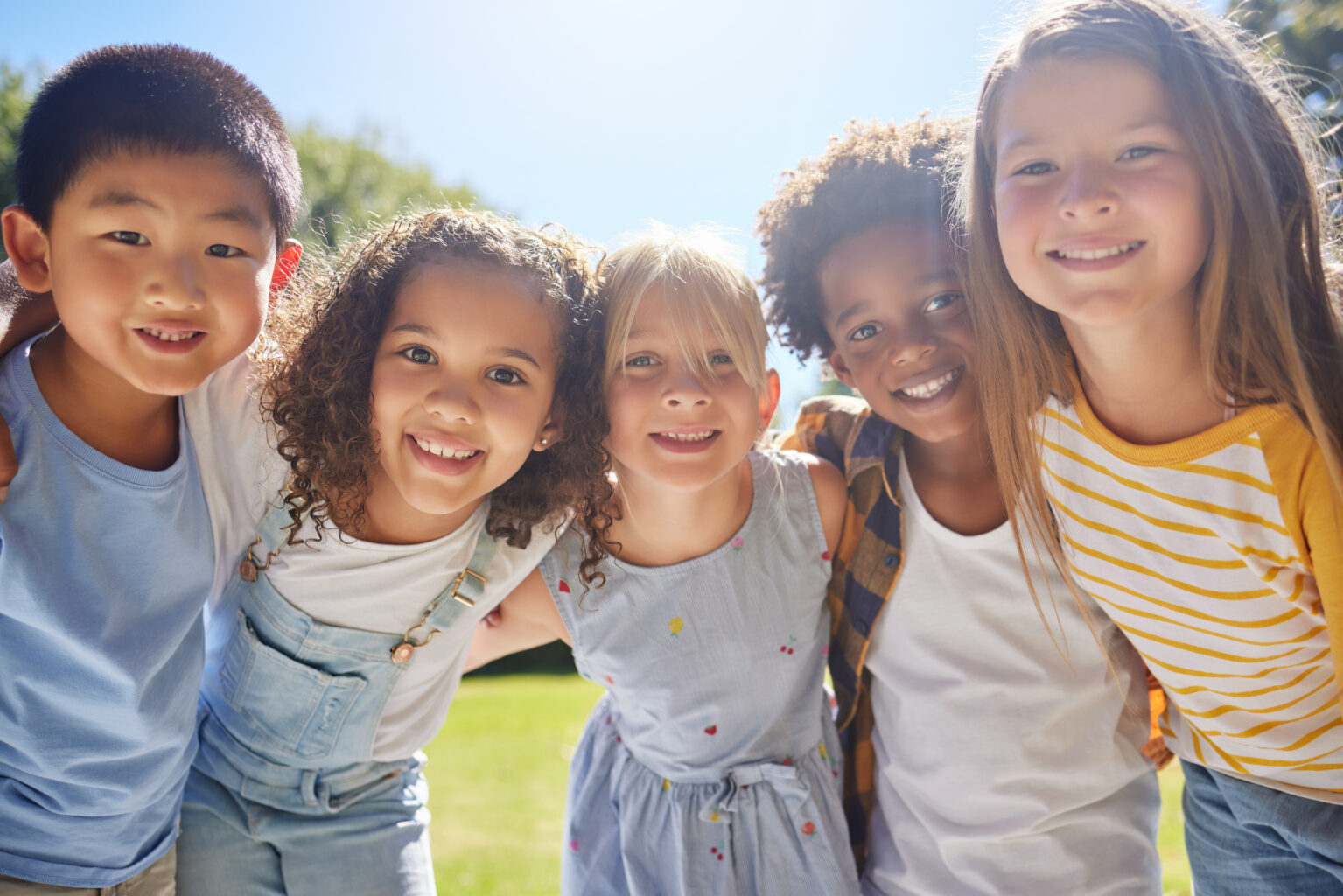 Five diverse children standing close together outdoors on a sunny day, all smiling warmly at the camera with their arms around each other's shoulders.