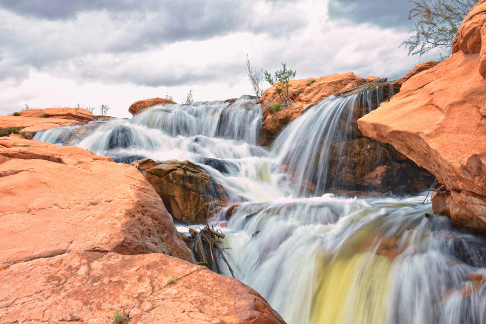 Multiple waterfalls cascade down red sandstone rock formations creating tiered pools with flowing white water against orange-colored stone under a cloudy sky.