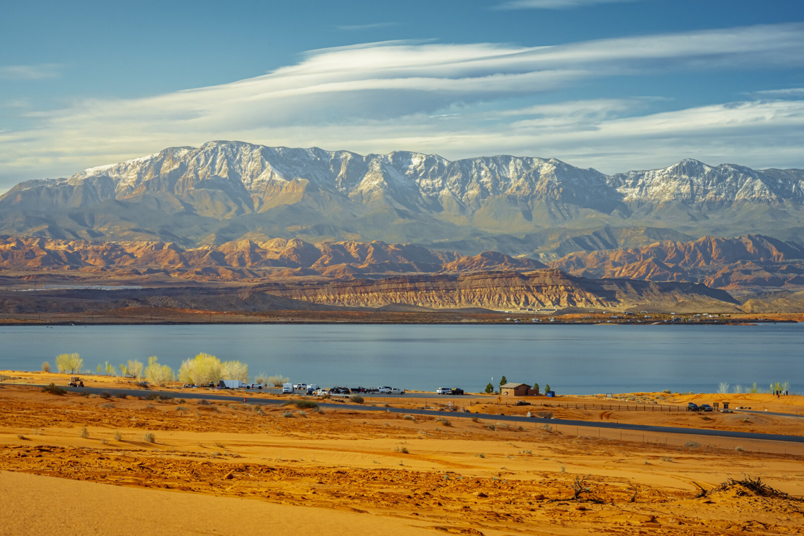 Expansive desert landscape with golden terrain in foreground, large blue lake in middle distance, and dramatic snow-capped mountain range under cloudy blue sky.