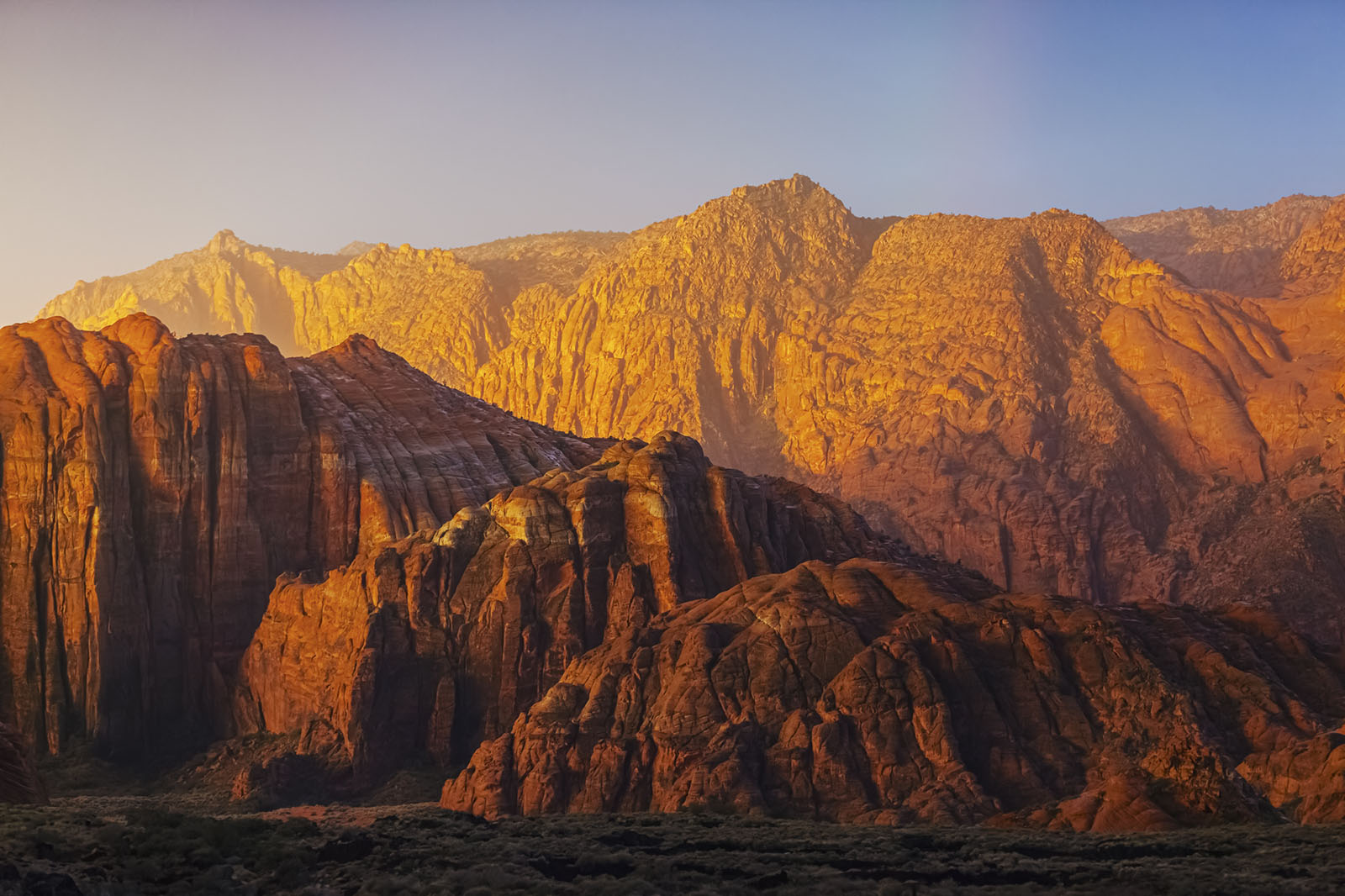 Dramatic golden sunset light illuminates towering red sandstone cliffs and rocky formations in a desert canyon landscape with layered geological features.