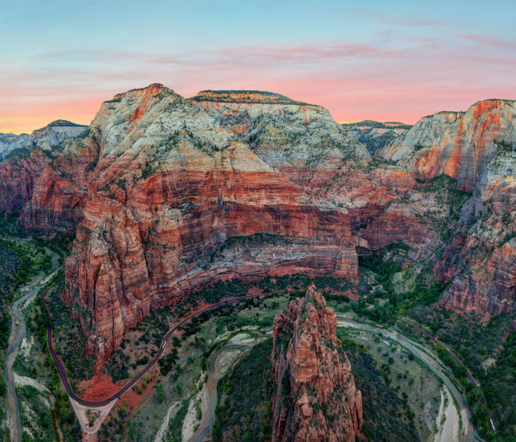 Aerial view of dramatic red and orange sandstone canyon formations with layered rock cliffs surrounding a winding river valley during sunset with pink sky.