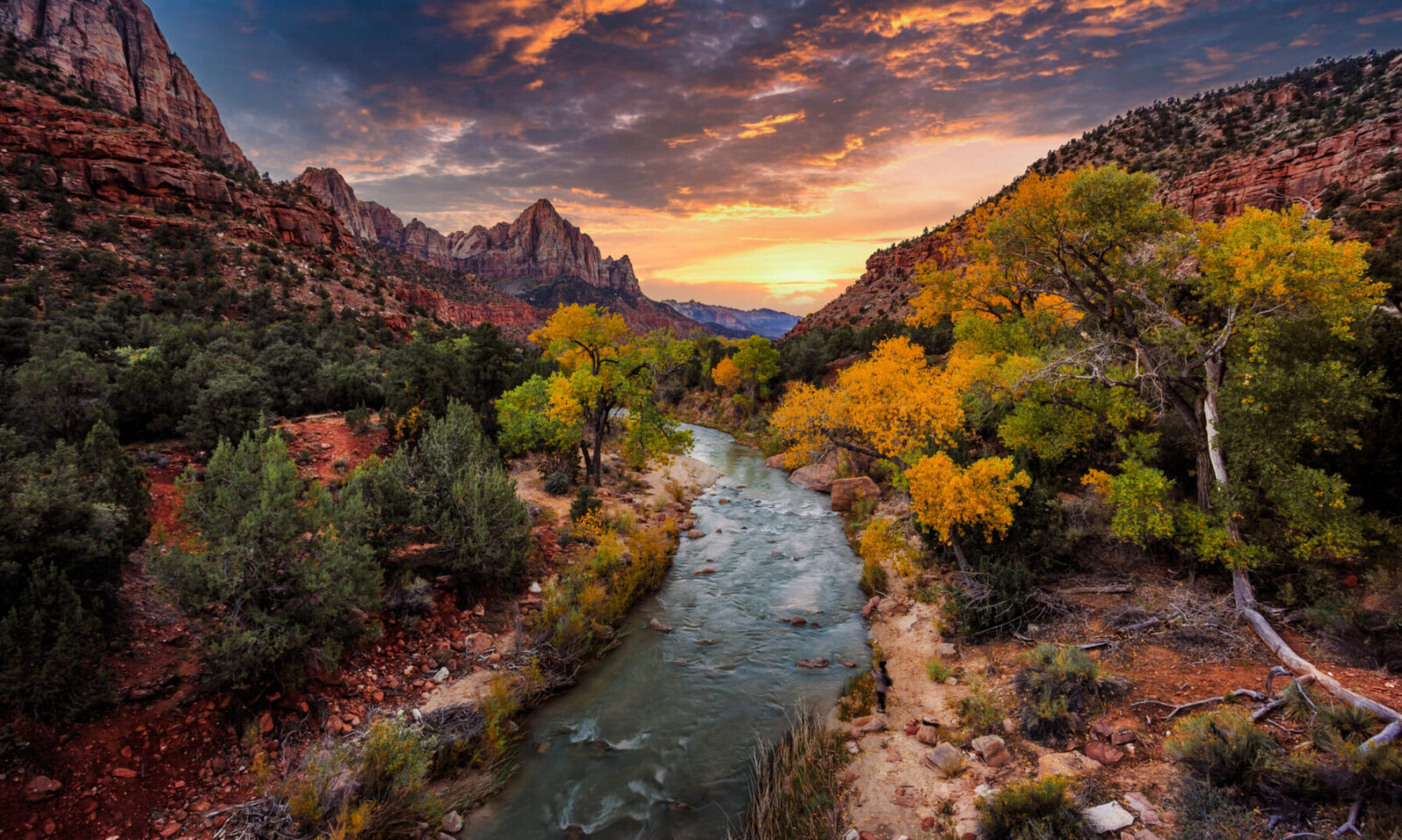 Winding river flows through canyon valley with towering red rock formations under dramatic sunset sky while autumn trees display golden and orange foliage along riverbanks.