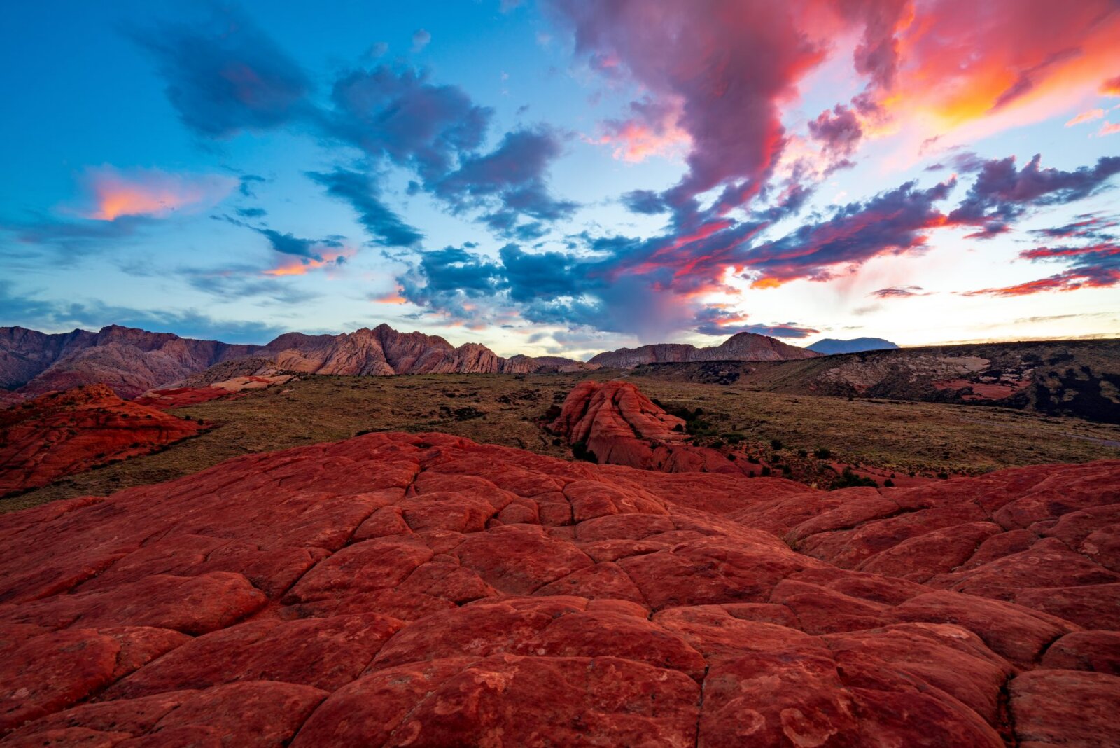 Dramatic sunset sky with vibrant blue and pink clouds above red sandstone rock formations in a desert landscape with layered stone terrain.