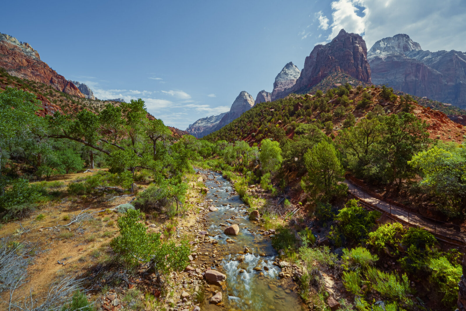A clear stream winds through a lush green valley surrounded by dramatic red and white sandstone cliffs under a bright blue sky with wispy clouds.