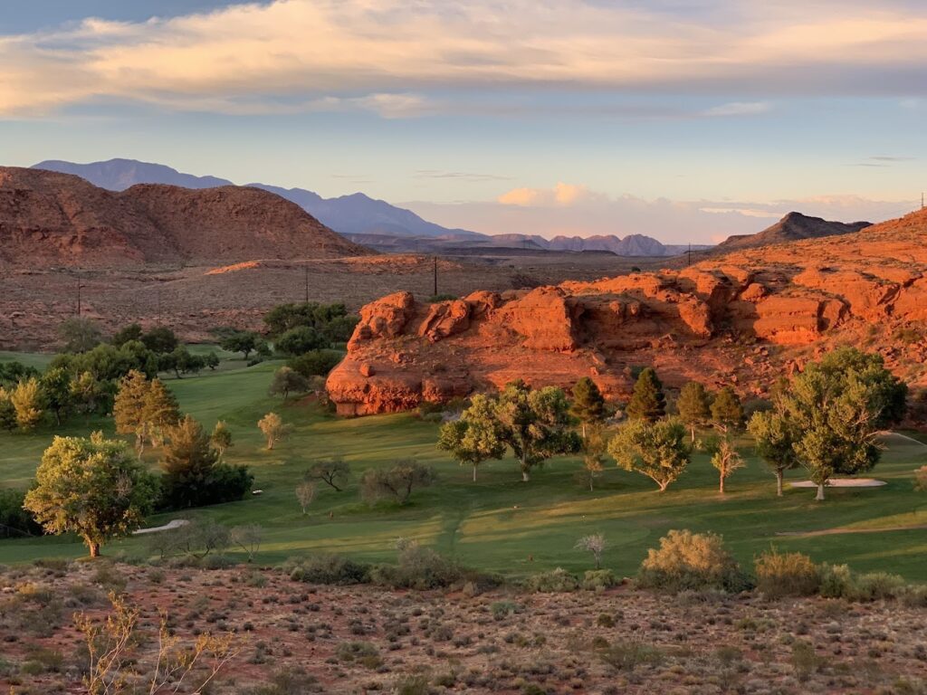 A scenic view of Dixie Red Hills Golf Course near Settlers Point RV Park in Utah, featuring lush greens and dramatic red rock formations at sunset.
