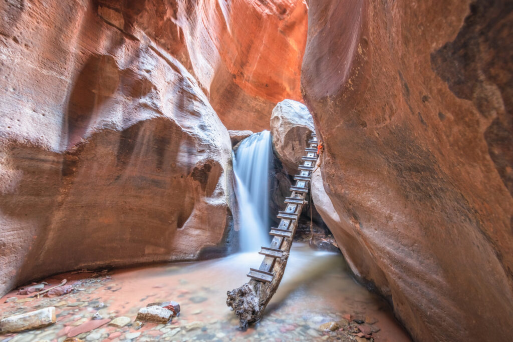 Narrow slot canyon with smooth sandstone walls in warm orange and pink tones, wooden ladder leading down through carved rock formations with water flowing below