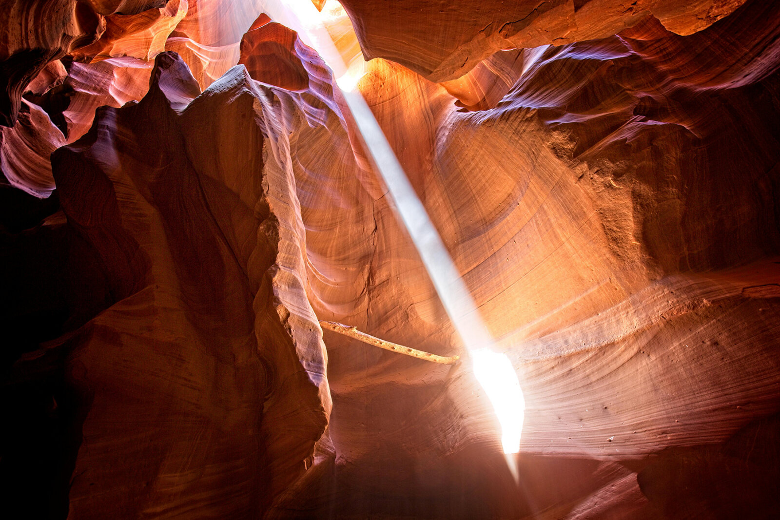 Brilliant beam of sunlight streaming down through opening of narrow slot canyon with smooth sculpted sandstone walls displaying rich orange and purple hues.