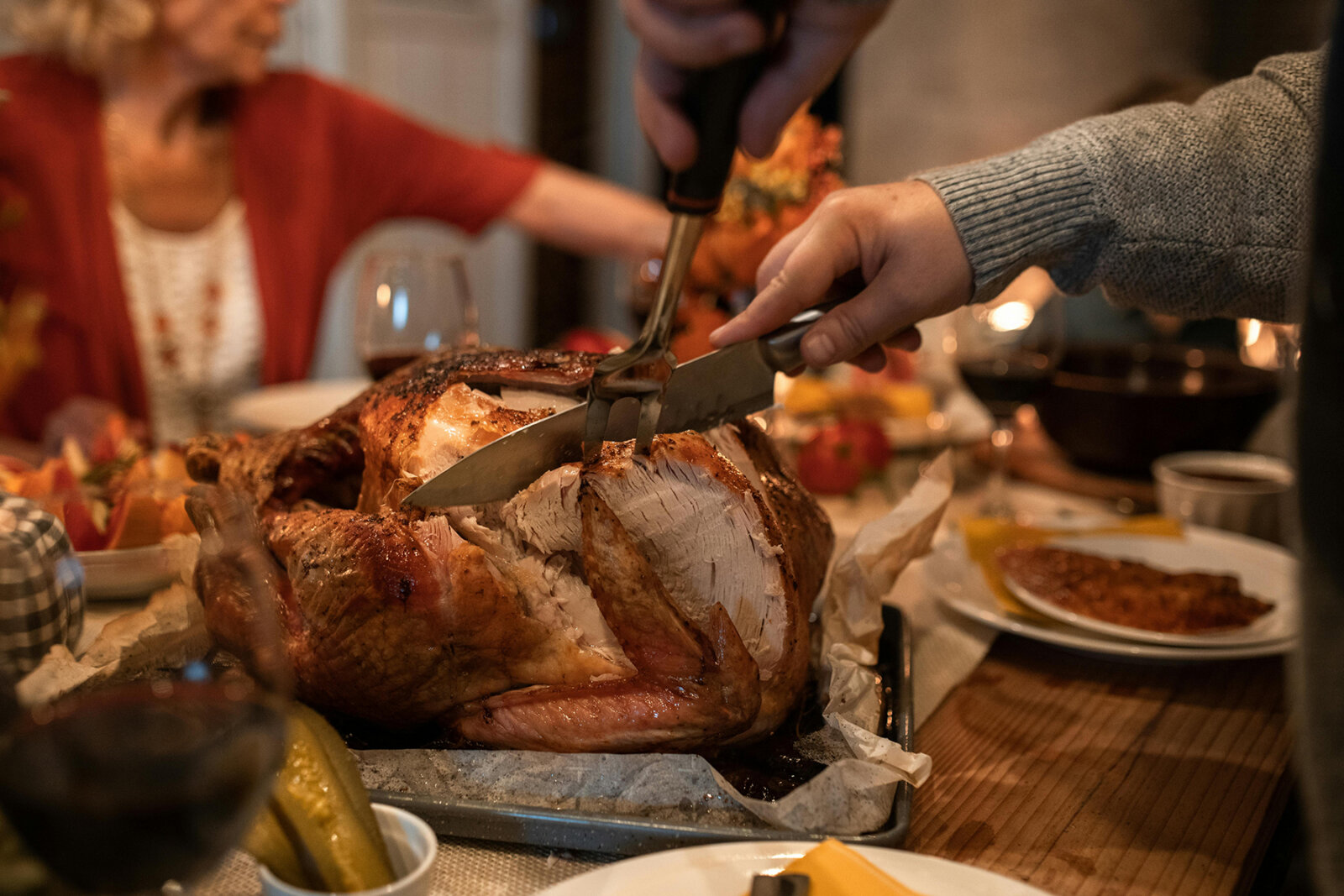 Hands using a carving knife to slice a golden-brown roasted turkey on a dinner table with wine glasses and plates in the background.
