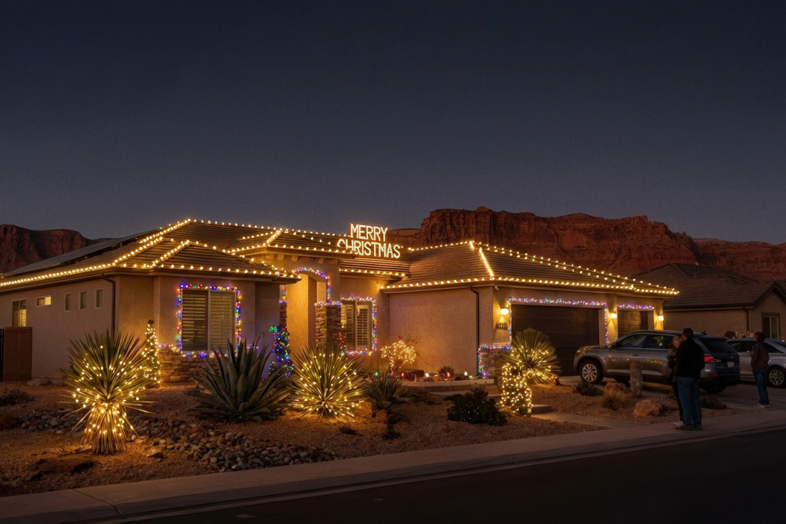 Single-story desert home decorated with warm white Christmas lights along the roofline and wrapped around desert plants in the front yard at dusk.
