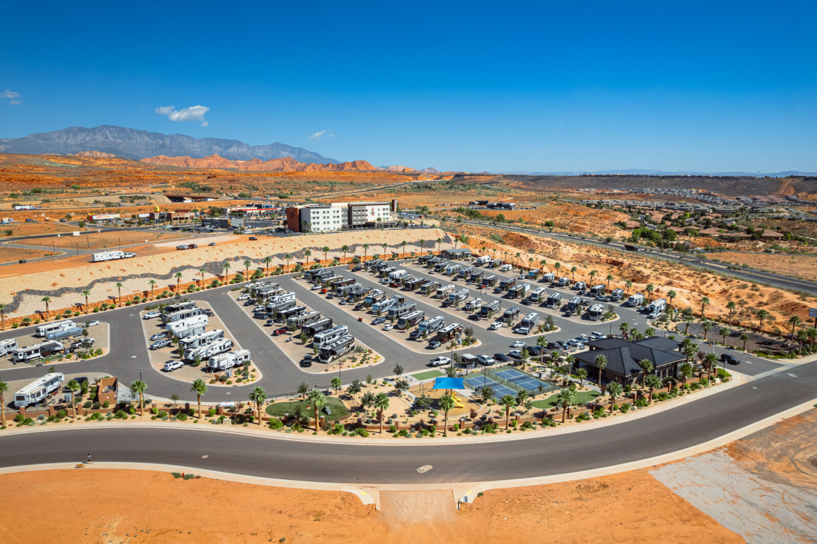 Aerial view of a recreational vehicle park with numerous RVs arranged in organized rows on paved roads, featuring a swimming pool area and surrounded by desert terrain under clear blue skies.