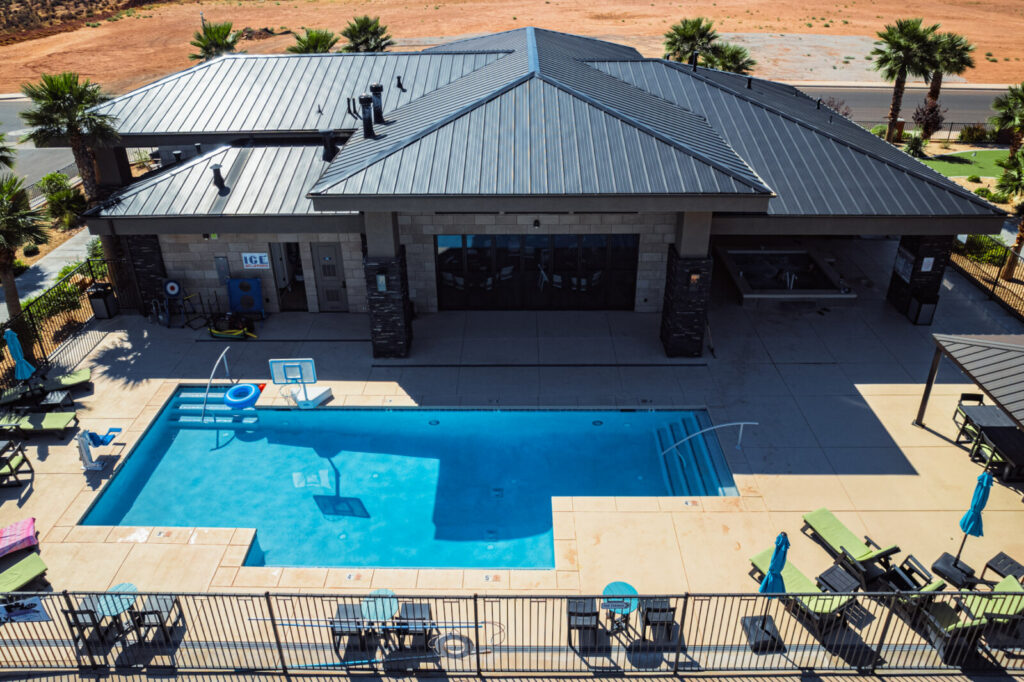 Aerial view of a modern single-story building with gray metal roof next to a large rectangular swimming pool surrounded by concrete decking and outdoor furniture.