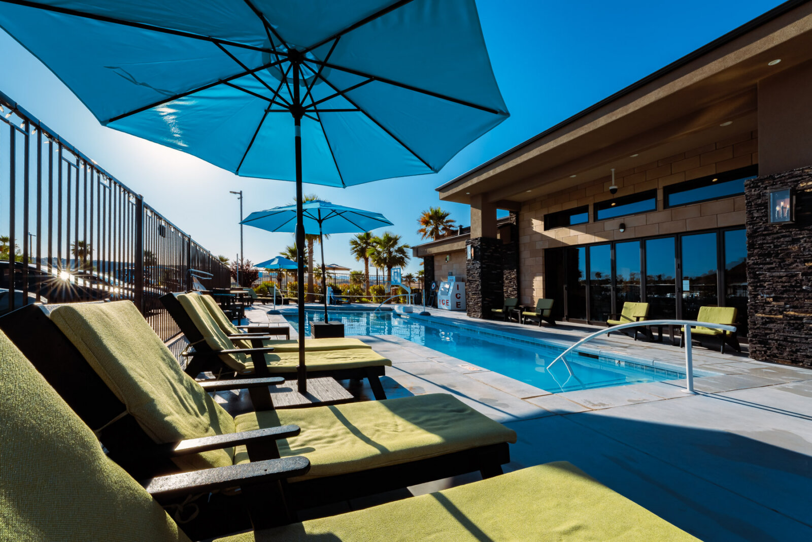 Outdoor pool deck featuring green upholstered lounge chairs arranged under large blue umbrellas with a modern clubhouse building and swimming pool in the background