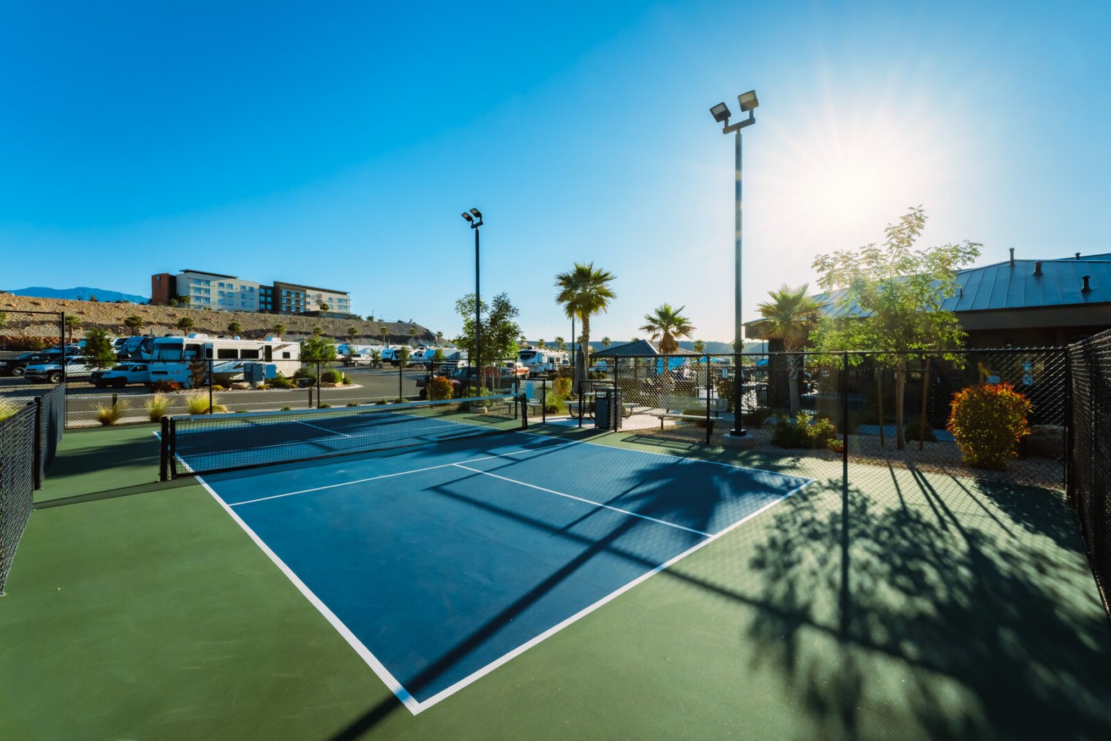 Outdoor tennis court with blue and green surface surrounded by black fencing, tall light poles, palm trees and landscaping under bright sunny blue sky.