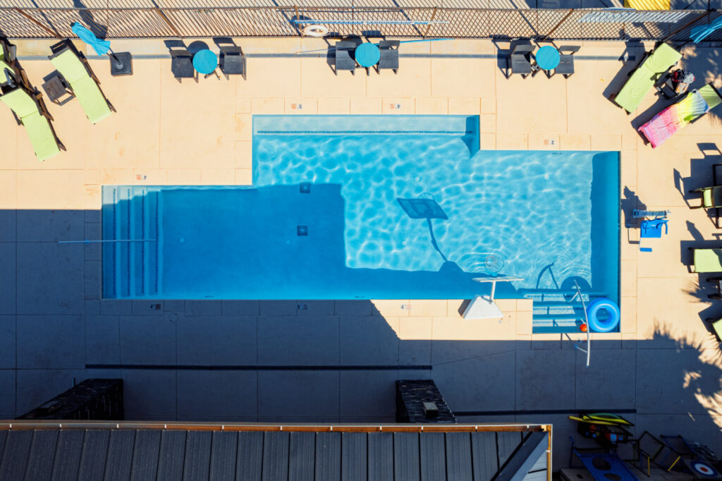 Aerial view of a rectangular swimming pool with crystal blue water surrounded by concrete decking, colorful lounge chairs, and umbrellas arranged around the perimeter.