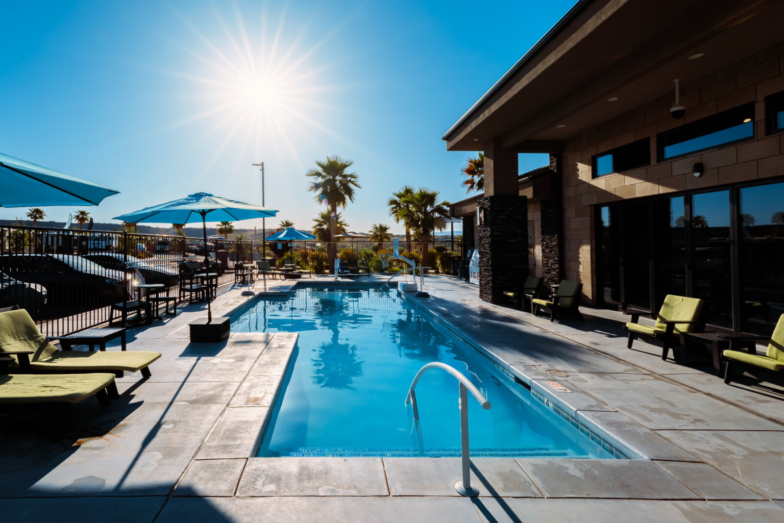 Sparkling blue swimming pool with handrail surrounded by stone decking, modern clubhouse building with large windows, palm trees, colorful lounge chairs and umbrellas under bright sunshine.