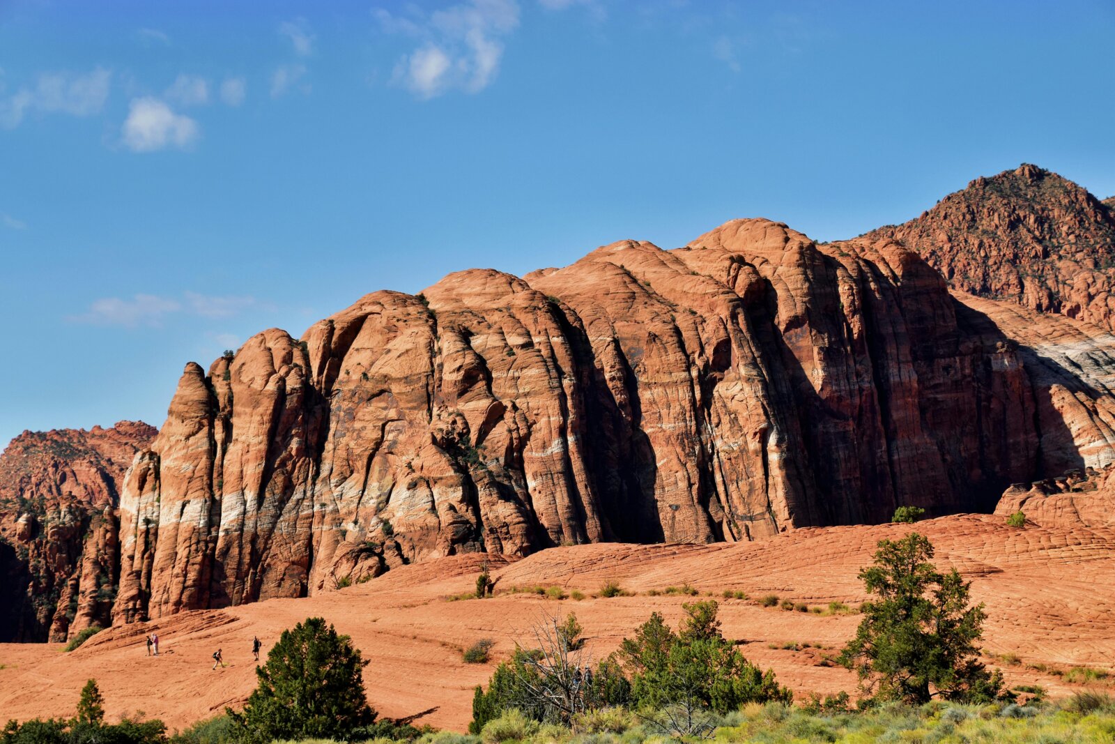 Majestic red rock formations reaching up towards a clear blue sky with fluffy white clouds, surrounded by a rugged desert landscape. | Image by Jeffrey Eisen