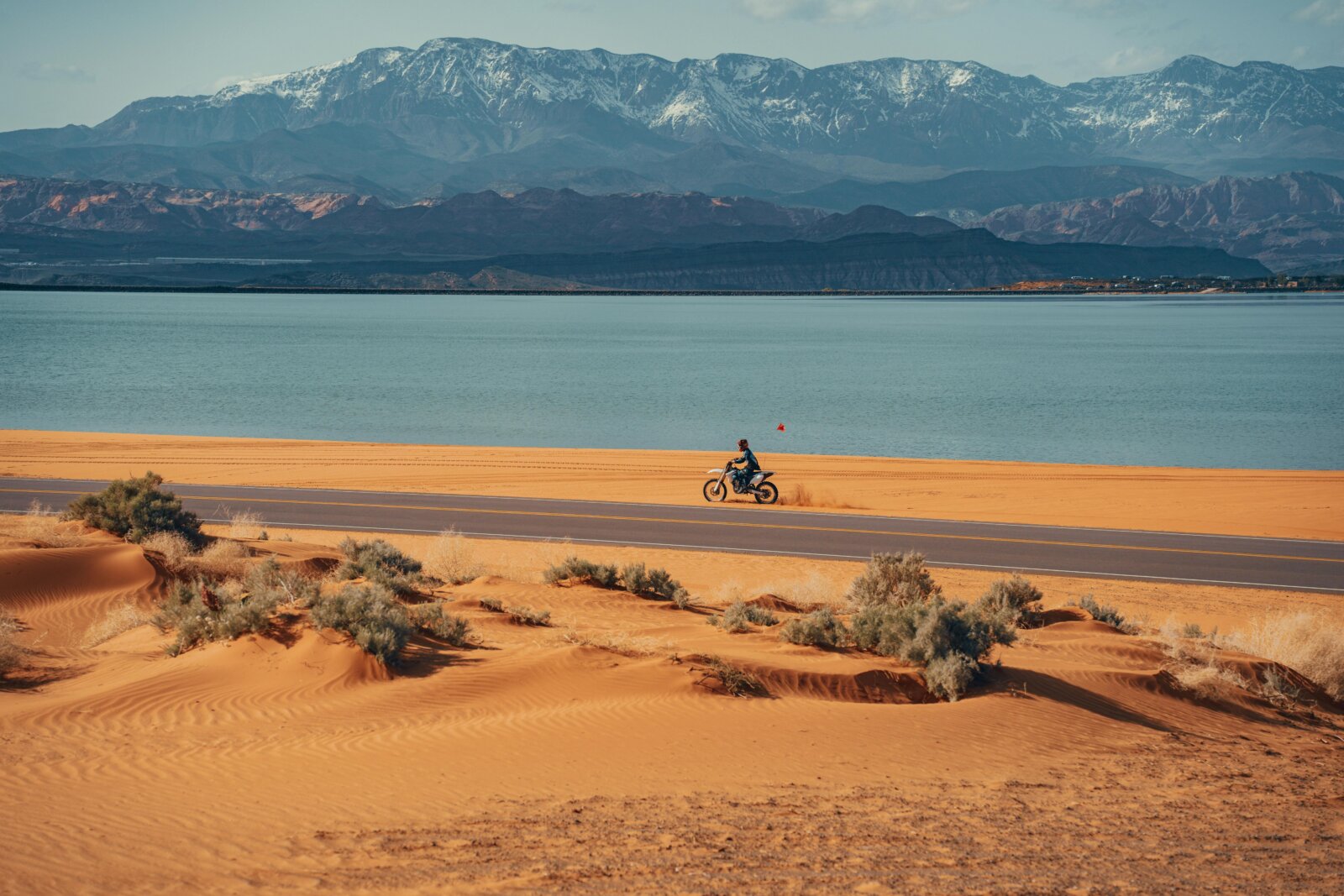 A motorcyclist rides along a paved road between orange sand dunes and a large blue lake with snow-capped mountains in the background.