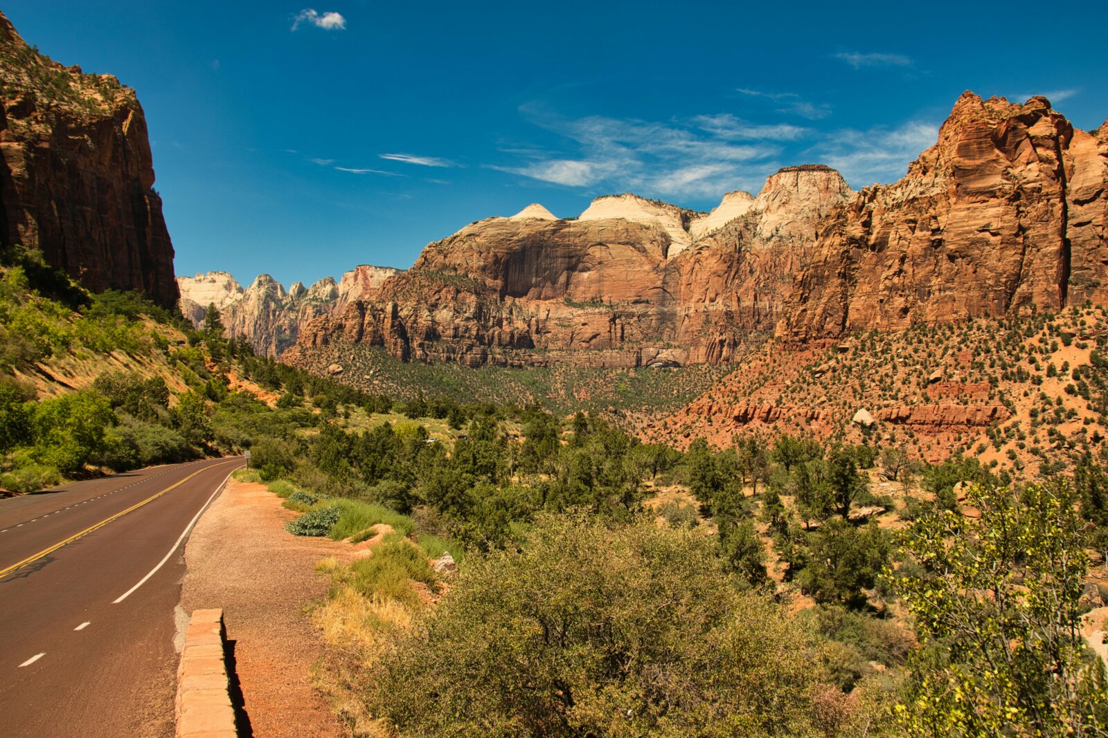 A breathtaking desert landscape with towering rocky cliffs, a winding road leading to a scenic overlook, and a vibrant blue sky with wispy white clouds. | Image by Gijs Jakobs