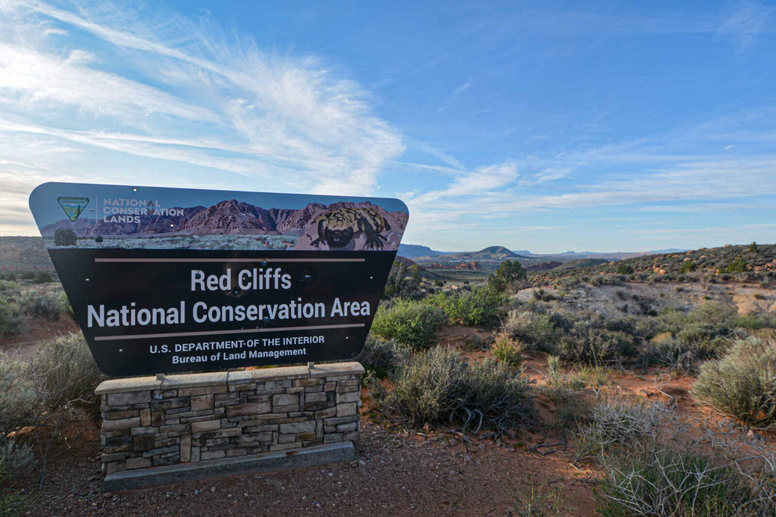 Large stone sign marking the entrance to the Red Cliffs National Conservation Area, with rocky desert terrain and a blue sky with wispy clouds in the background.
