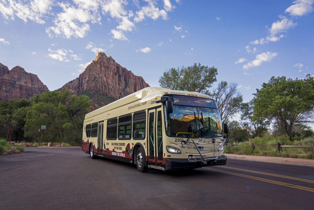 A cream and red shuttle bus drives along a road with tall red rock formations and trees in the background.