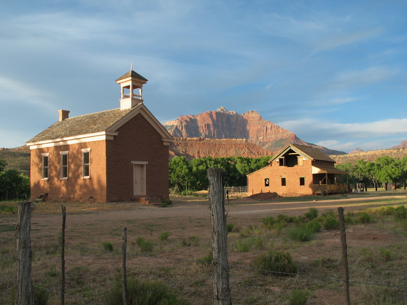 A historic brick schoolhouse with a bell tower stands before dramatic red rock cliffs under a blue sky.