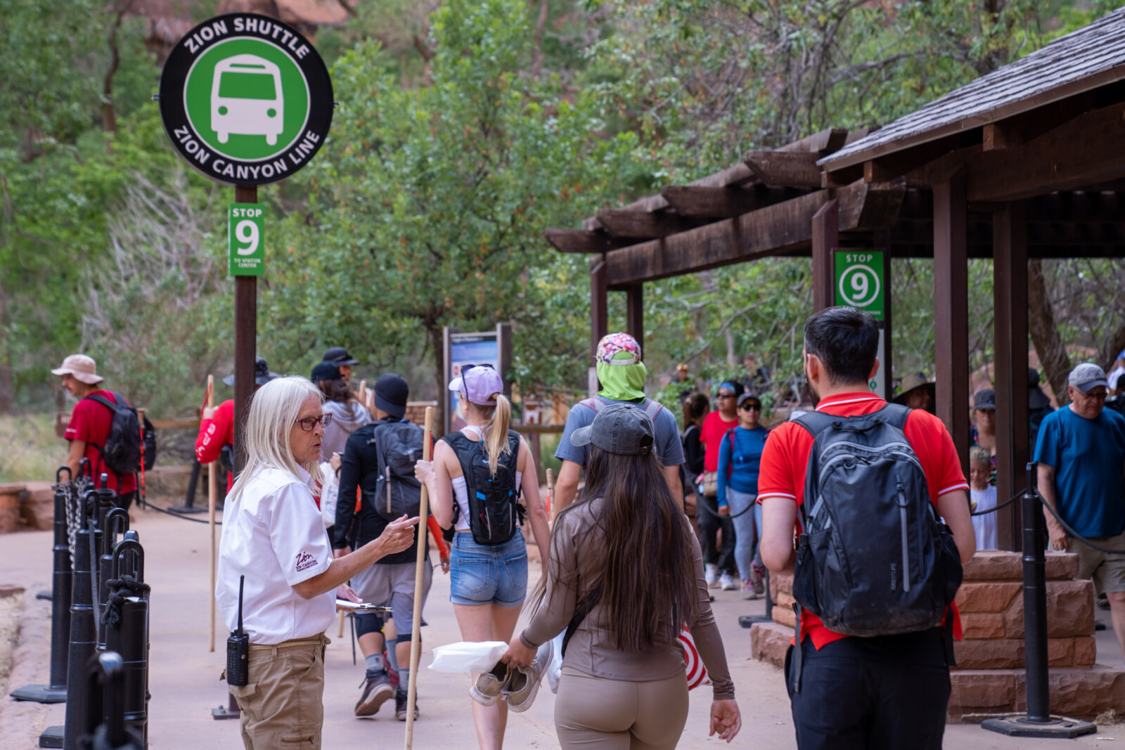 A crowd of visitors walking toward a shuttle bus stop sign along a wooded path.