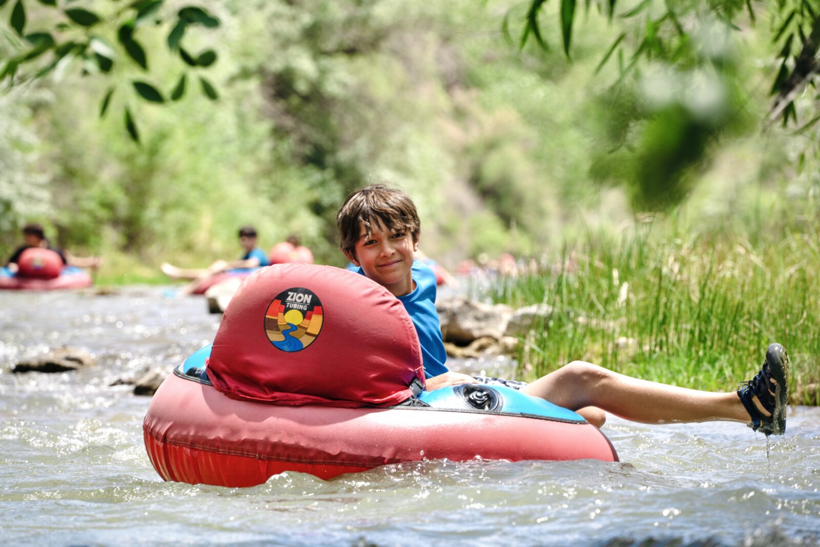 A smiling boy relaxes in a red inflatable tube floating down a shallow rocky river.