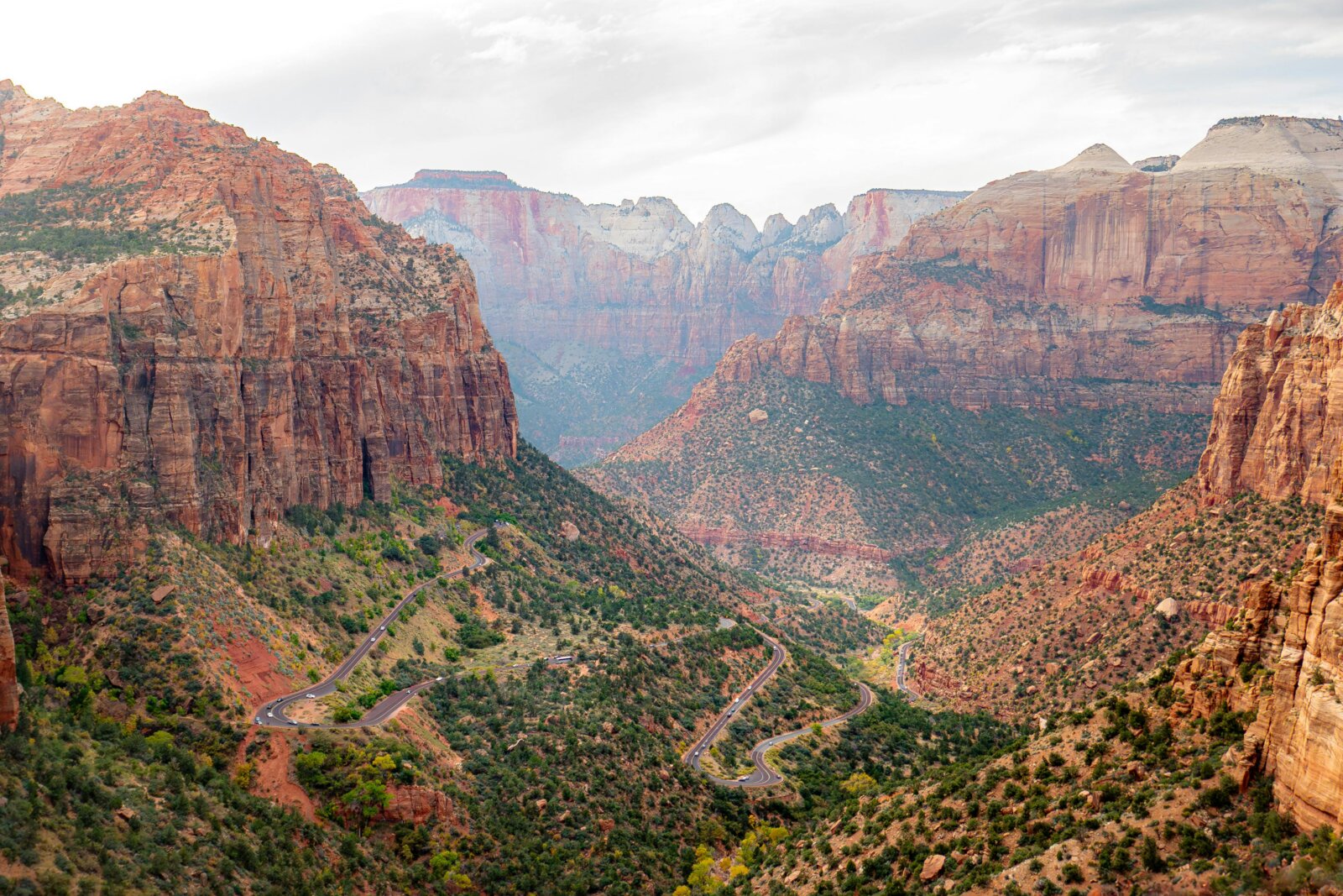 A winding road curves through a deep rocky canyon valley flanked by towering red sandstone cliffs and green vegetation.