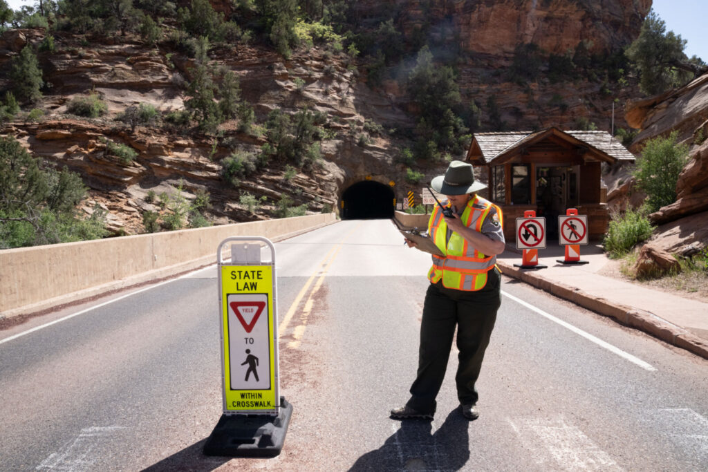 A park ranger in a safety vest uses a radio while standing on a road near a tunnel entrance.