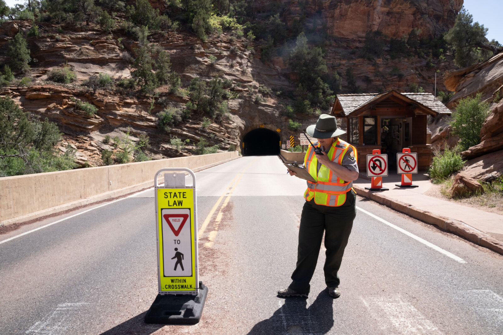 A park ranger in a safety vest uses a radio while standing on a road near a tunnel entrance.