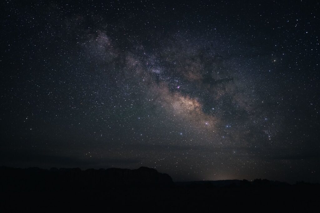 The Milky Way galaxy arcs across a dark night sky above a silhouetted landscape with rolling hills.