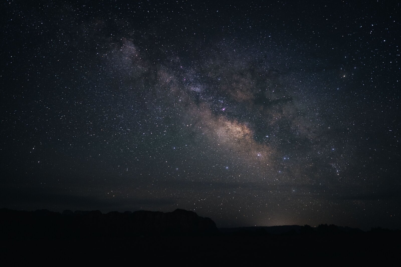 The Milky Way galaxy arcs across a dark night sky above a silhouetted landscape with rolling hills.