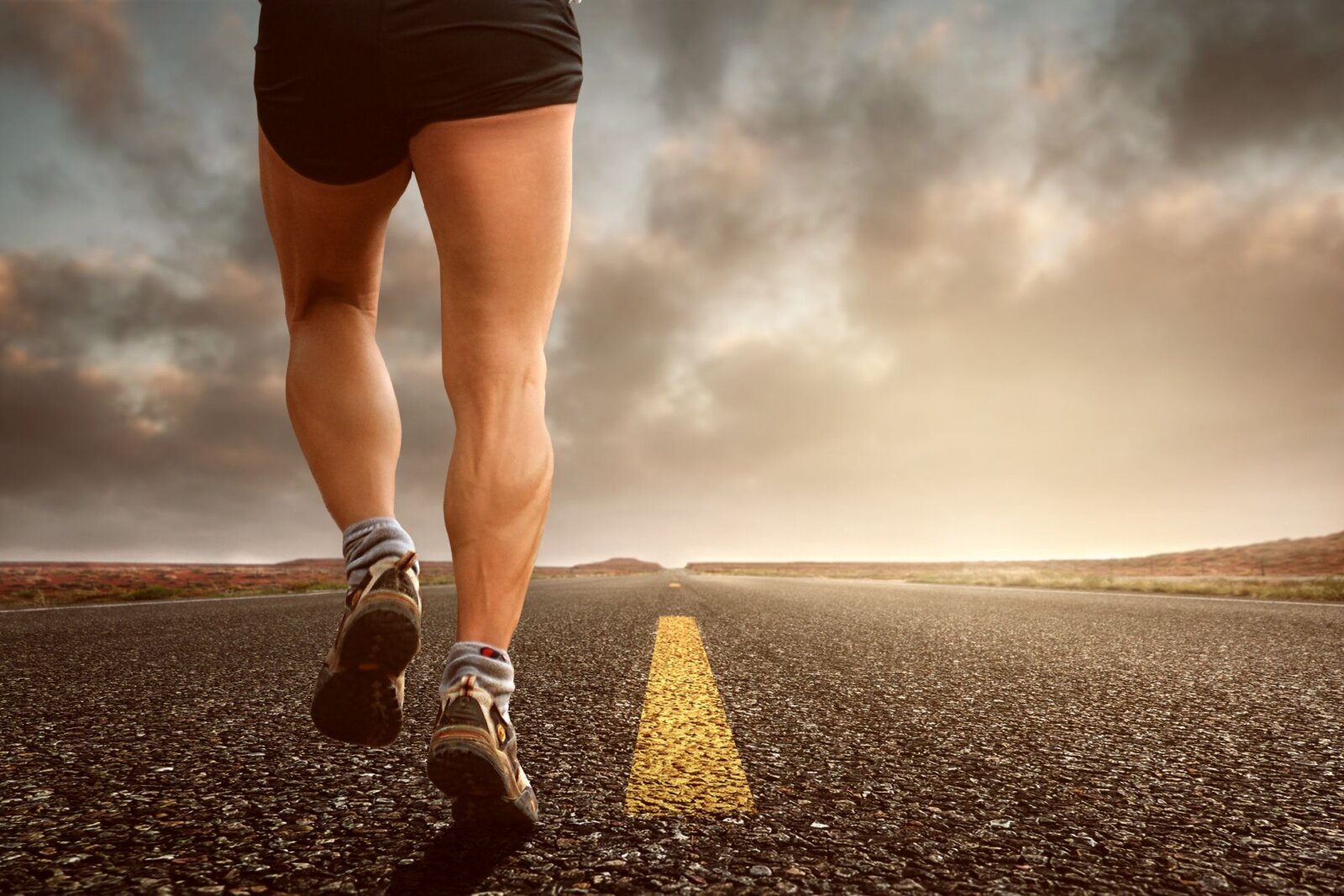 A runner's muscular legs stride along an open road beneath a dramatic cloudy sky at sunset.